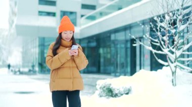 Güzel mutlu Asyalı genç kız ya da öğrenci soğuk havada kışın karlı sokaklarda yürürken cep telefonu kullanıyor. Gülümse. Akıllı telefonları olan genç bir kadın mesajlaşarak kayıyor. Dışarıda, dışarıda.