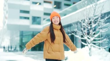 Soğuk kış havasında şehrin sokaklarında dans eden neşeli ve neşeli genç bir kadın. Mutluluk. Asyalı kız öğrenci, modern bir şehir geçmişinde gülümseyerek yürüyerek zıplıyor. Sevinç, mutluluk