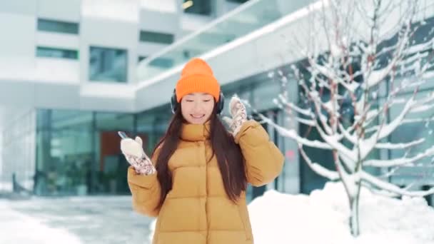 아름다운 아시아 여성은 눈내리는 헤드폰을 음악을 온라인 사용하며 날씨가 소녀가 — 비디오
