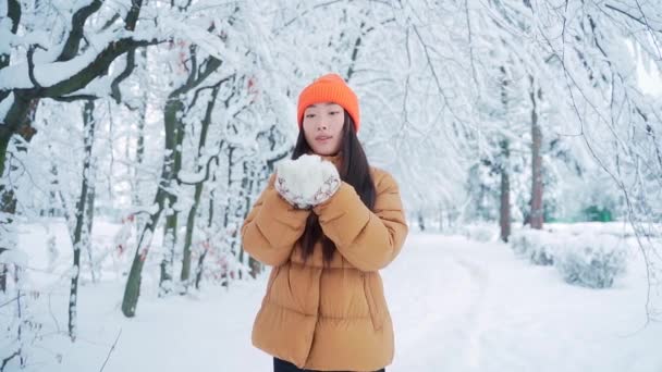 美しい冬のアジアの女の子は霜の多い冬の公園で雪を吹いています 屋外だ スノーフレークを飛んで 寒い屋外で楽しんでる若い女性 幸せな陽気な魅力的な美しい女性の笑顔 楽しみ方 — ストック動画