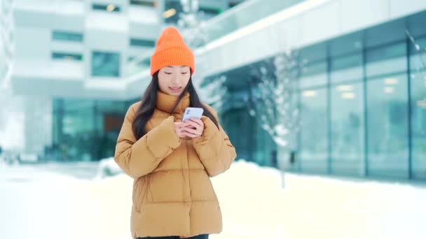 美丽而快乐的亚洲少女或学生在寒冷的冬季雪城街道上散步时使用手机 笑一个手里拿着智能手机的年轻女人在滚动发短信 — 图库视频影像