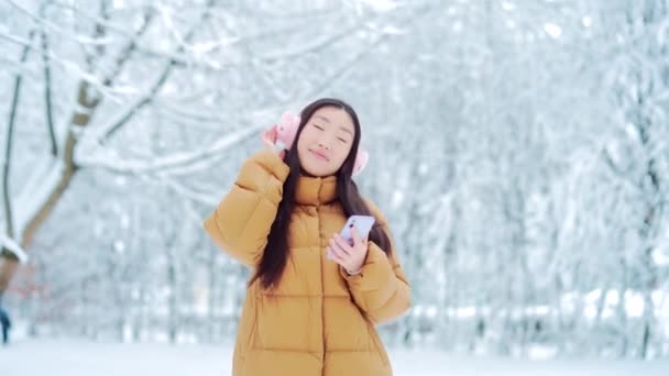 雪の冬の日に音楽を聞いて公園を歩く幸せな陽気な面白いアジアの十代の女の子の学生 携帯電話のスマートフォンを持つ若い女性は寒い天候で踊ることを楽しむ 外の屋外 — ストック動画