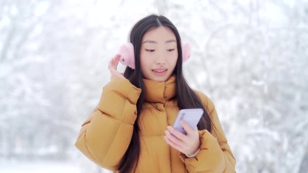 在一个雪天的公园里散步 听着音乐 快乐而有趣的亚洲少女学生 拿着手机的年轻女人喜欢在寒冷的天气跳舞 — 图库视频影像