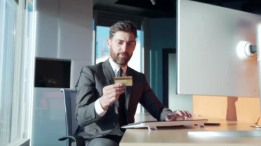Banka ödemesi yapan mutlu bir iş adamı, modern ofisteki bilgisayarın başında duran bir kredi kartı tutuyor. Masadaki takım elbiseli erkek girişimci faturaları ödemek için internetten alışveriş yapıyor. Altın müşteri ödüyor.