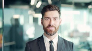 Modern cam ofiste dikilip kameraya bakan resmi takım elbiseli kendine güvenen iş adamının portresi. Sakallı yakışıklı beyaz erkek işadamı gülümsüyor. İçeride mutlu ve neşeli bir insan. Kapat.