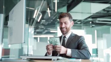 Modern ofis işyerinde oturan başarılı iş adamı nakit para sayıyor. Erkek girişimci ya da çalışan, uygulamayla çevrimiçi kazanılan dolarları hesaplar.