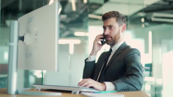モダンなオフィスのデスクに座ってる携帯電話の男だ 白人男性ビジネスマンがスーツを着ている 従業員起業家として成功を収め 携帯電話でコミュニケーションを取り — ストック動画