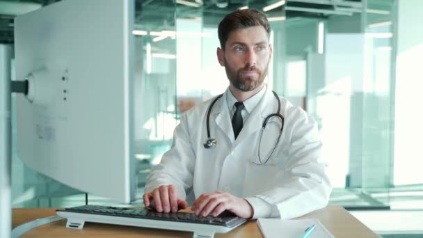自信のあるひげそりの成熟した男性医師薬剤師またはセラピストは デスクワークで座っている病院のオフィスでコンピュータで作業し 無線を入力します 患者とのオンライン対応を規定しています — ストック動画