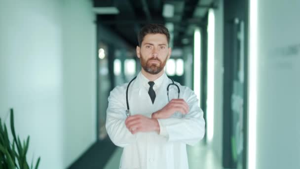 現代の病院の診療所を背景にカメラを真剣に見つめる自信に満ちた白人医師の肖像画 男性セラピストや医師は 屋内で腕を組んで廊下に立っている — ストック動画