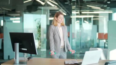 Modern ofiste, bilgisayarın önünde, dambıllarla egzersiz yapan kadın ofis çalışanı. İşyeri Sağlık Hizmetleri 'nde çalışan iş kadını fitness yöneticisi. Sağlıklı bir yaşam tarzını bozmak. Çevrimiçi