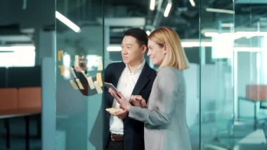 Ofiste takım çalışması toplantısı. İki mutlu Asyalı iş arkadaşı yazı yazıyor. Yapışkan notlar. Çalışma örgütünün strateji planını tartışan cam duvar tahtasına bakın.