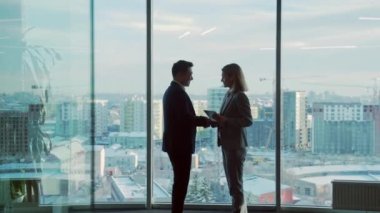 Modern ofis odasında iş ortaklarıyla tanışma silueti pencere arkasında duruyor. Kapalı alanda. Asyalı adam bir kadınla tokalaşıyor. İki işadamı müzakere hakkında konuşuyor.