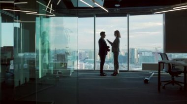 Modern ofis odasında iş ortaklarıyla tanışma silueti pencere arkasında duruyor. Kapalı alanda. Asyalı adam bir kadınla tokalaşıyor. İki işadamı müzakere hakkında konuşuyor.