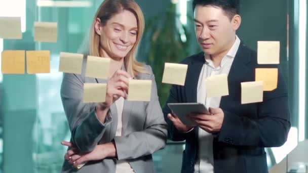 オフィスでのチームワークの会議 2つの幸せなビジネスアジアの同僚のチームは それは粘着ノートをポストに書き込みます作業組織のタスク管理にガラス壁スクラムボードブレインストーミングを見て戦略計画を議論 — ストック動画