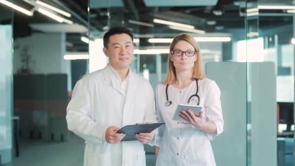 病院でカメラを見ている2人の笑顔の幸せな医者を描いたものです アジア系の男性と白人女性をグループ化する チーム多様な男性看護師と女性看護師クリニックヘルスケアに立つ医療スタッフ — ストック動画