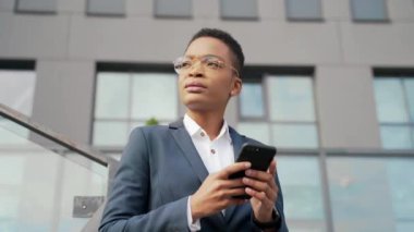 Kendine güvenen Afro-Amerikan kadın modern ofis merkezinin yanında duran cep telefonu akıllı telefonu kullanıyor. Dışarıdaki resmi takım elbiseli siyahi kadın girişimci mesajları okuyor.