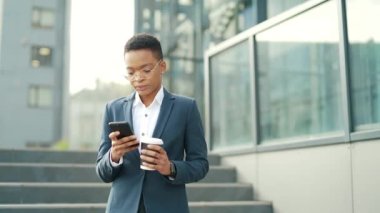 Kendine güvenen Afro-Amerikan kadın modern ofis merkezinin yanında duran cep telefonu akıllı telefonu kullanıyor. Dışarıdaki resmi takım elbiseli siyahi kadın girişimci mesajları okuyor.