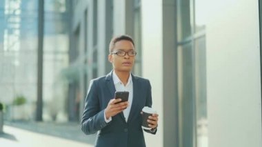 Şehir merkezinde elinde cep telefonu ve fincan kahveyle yürüyen siyahi bir iş kadını. Afro-Amerikalı kadın girişimci şehir geçmişinde akıllı telefon kullanıyor. kendine güvenen ofis çalışanı