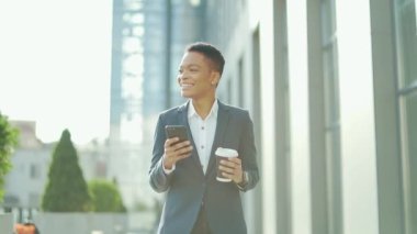 Mutlu gülümseyen zenci kadın cep telefonu ve fincan kahveyle şehir caddesinde yürüyor. Afro-Amerikalı kadın girişimci şehir geçmişinde akıllı telefon kullanıyor. Memnun ofis çalışanı