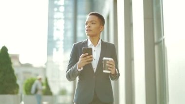 Şehir merkezinde elinde cep telefonu ve fincan kahveyle yürüyen siyahi bir iş kadını. Afro-Amerikalı kadın girişimci şehir geçmişinde akıllı telefon kullanıyor. kendine güvenen ofis çalışanı