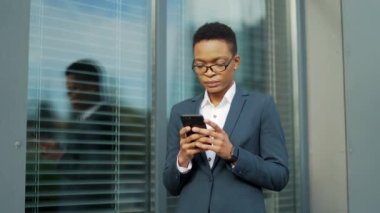 Kendine güvenen Afro-Amerikan kadın modern ofis merkezinin yanında duran cep telefonu akıllı telefonu kullanıyor. Dışarıdaki resmi takım elbiseli siyahi kadın girişimci mesajları okuyor.