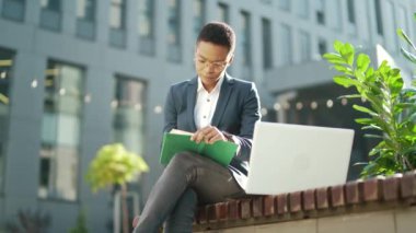 Genç Afro-Amerikan iş kadını öğrenci kenar mahallede bir bankta dizüstü bilgisayar ve defter ile oturuyor ve açık hava üzerine çalışıyor. Gözlüklü serbest çalışan kadın, sokak e-öğreniminde uzaktan öğreniyor.