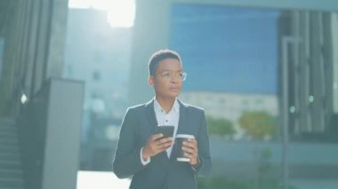 Kendine güvenen başarılı genç iş kadını cep telefonuyla modern ofis binasına karşı hareket ediyor şehir merkezindeki iş kadını elinde bir kahve fincanıyla cep telefonu akıllı telefonu kullanıyor.
