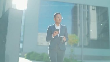 Kendine güvenen başarılı genç iş kadını cep telefonuyla modern ofis binasına karşı hareket ediyor şehir merkezindeki iş kadını elinde bir kahve fincanıyla cep telefonu akıllı telefonu kullanıyor.