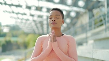 Genç Afro-Amerikan fitness kadını modern bir stadyumun minderinde yoga yapıyor. Açık havada. Siyah dişi, Lotus pozisyonunda meditasyon yapar ve rahatlar. Derin nefes alır ve parktaki gözleri kapalıdır.