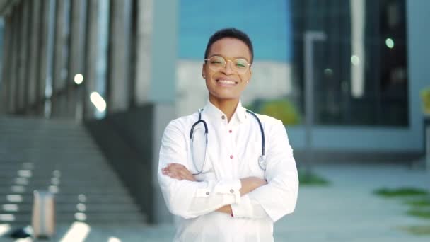 アフリカ系アメリカ人の医師が腕を組んでカメラを見て笑顔で肖像画を閉じると 外の現代的な病院を横切った 幸せな医療を信じています 診療所近くのスクラブを着た女性科学者療法士 — ストック動画