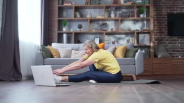 senior active gray-haired woman doing exercise, fitness aerobics, stretching and gymnastics. online workout lessons using laptop sitting on the mat at home indoors. Mature female healthy lifestyle