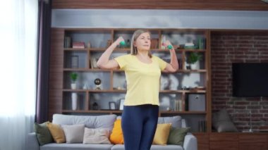 Sportif, yaşlı, yaşlı, kır saçlı kadın evinde elinde dambıllarla egzersiz yapıyor. Yaşlı, olgun bir kadın içeride spor yapıyor. İnsanlar hakkında fikir, evde spor, spor