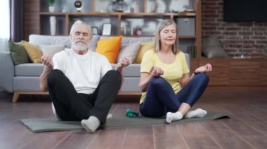 Üst sınıf çifti evde yoga yapıyor. Kapalı alanda. Yetişkin bir aile oturma odasında yoga minderinde nefes egzersizi yapıyor. Meditasyon yap ve gözlerin kapalı rahatla. Lotus Pozu 'nda sakinleş. Emeklilik