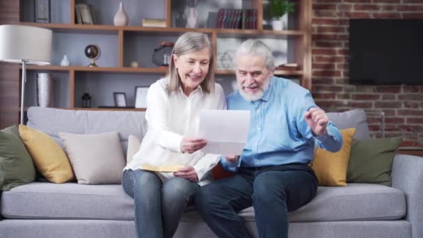 年长的灰白头发的家庭成员坐在家里高兴地收到信件或文件 兴高采烈的丈夫和妻子退休了 一边笑着一边看这个好消息 老年人在看着 快乐的祖父母 — 图库视频影像