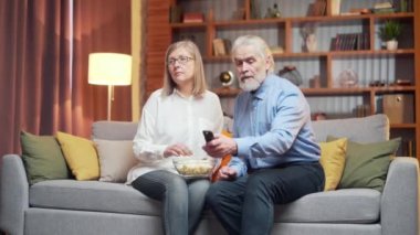 Gri saçlı yaşlı çift televizyon izlemek ve uzaktan kumandayla kanal değiştirmek için bir televizyon programı seçiyor. Yaşlı, olgun bir aile babası, karı koca bir dizi seçemez. Haberleri ya da filmleri izlemek