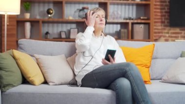 Evdeki kanepede müzik dinleyen son sınıf öğrencisi bir kadın. Mutlu yaşlı kadın kulaklık takıyor. Akıllı telefonu tutuyor. Rahat ol, gözleri kapalı. Gri saçlı bayan rahatla meditasyon yap ve sakinleş. ses tedavisi