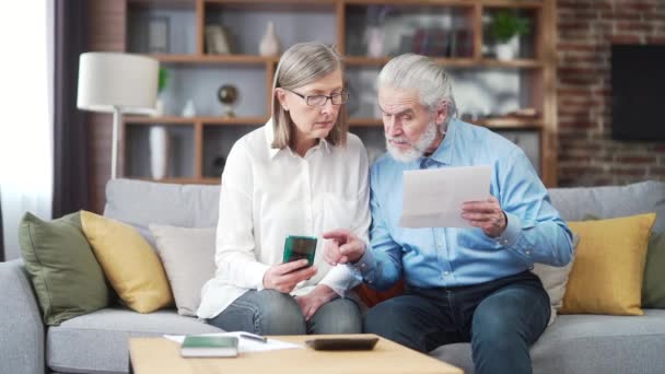 シニアファミリーカップル 白髪の老人とサービスの女性は アプリケーションの携帯電話を使用してデータ文書を入力します 自宅で退職者 公共料金や小切手はコンピューターのスマートフォンを使って — ストック動画