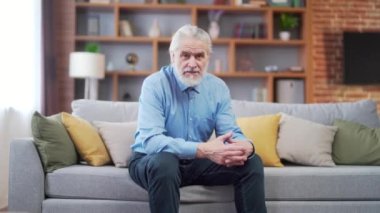 Portrait of a confident gray-haired senior mature man looking at camera. sitting in the living room on the couch at home. Indoor. Headshot of charismatic senior male on cosy sofa