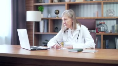 Modern hastane kliniğinin masasında çalışan olgun kıdemli doktor kadın. Bilgisayar kullanıyor, reçeteli oturma izni yazıyor. kadın pratisyen eczacı bilgisayardan tıbbi notlar alıyor.