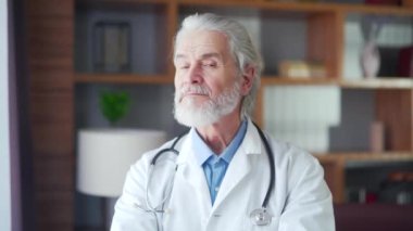 Ofisindeki modern bir hastane kliniğinde kameraya bakarak duran yaşlı, gri saçlı, deneyimli kıdemli doktor portresi. Kollarını kavuşturdu. Kendinden emin yaşlı hekim pratisyen doktor gülümsüyor.