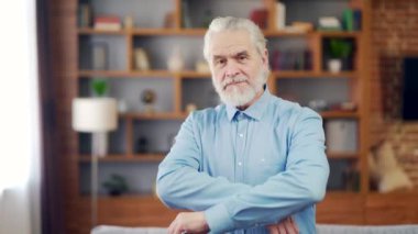 portrait senior gray-haired man with a beard looking at camera with his arms crossed. Indoors at home or a classic office. Business old retired male. Confident happy cheerful and smiling in the room