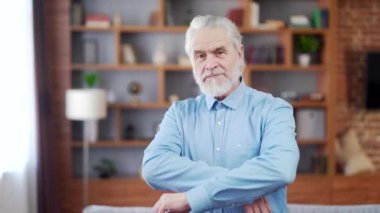 portrait senior gray-haired man with a beard looking at camera with his arms crossed. Indoors at home or a classic office. Business old retired male. Confident happy cheerful and smiling in the room