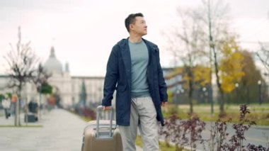 Asyalı erkek turist çantalı ya da valizini şehir caddesinde taşıyor. Yeni bir Avrupa şehrine gelen bir adam kaldırımda kentsel bir arka planda yürüyor. Sıkı giyin. Seyahat, turizm