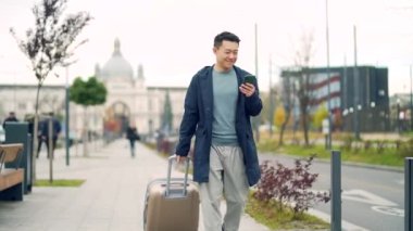 Asyalı erkek turist çantasıyla gidiyor ya da cep telefonu kullanarak bir bavulu şehir sokağına çekiyor. Yeni bir Avrupa şehrine gelen adam kaldırımda arka planda akıllı telefon kullanarak yürüyor. Dışarıda.