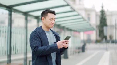 Asian man using phone at bus or tram stop. Asian people use smartphone waiting for bus station, orders a taxi application on the phone, book accommodation online for business travel