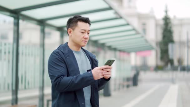 亚洲人在公共汽车或电车站使用电话 亚洲人使用智能手机等汽车站 在电话上订购出租车申请 在网上订房供商务旅行 — 图库视频影像