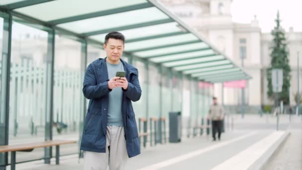 资深中国游客使用手机应用程序预订住宿和呼叫出租车 靠近机场的男子带着行李箱 亚洲人在巴士站用智能手机应用程序租车 等候公共交通工具 — 图库视频影像