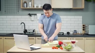 Asyalı adam mutfakta video dersi için hazırlanıyor. Bir adam internetten yemek pişirme dersi izliyor. Erkek, evde dizüstü bilgisayar kullanarak yemek hazırlıyor.