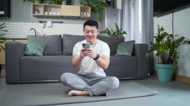 Asyalı adam oturma odasında lotus pozisyonunda yoga yapıyor. Meditasyon yapıyor ve içeride rahatlıyor. Fitness sporu aktif yaşam tarzı. Derslere bakmak için cep telefonu uygulaması kullanıyor.