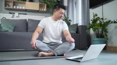Happy Asian evde yoga yapıyor, lotus pozisyonunda oturuyor, adam online dersler için dizüstü bilgisayar kullanıyor, aktif bir yaşam tarzı uyguluyor. dizüstü bilgisayar öğrenme derslerini izle